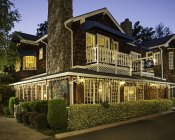 Exterior of inn at dusk with cedar shake siding and white trim, lit windows surrounded by trees and manicured shrubs