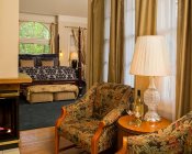 Two chairs with small table on wood floors near fireplace, and blue carpeted bedroom in background with arch top window