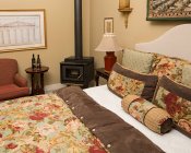 Bed with red, white, green, blue and brown floral comforter, tan walls, red chair with small table and wood stove