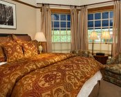 Bedroom with bay window, bed covered in red and gold pillows and comforter, two chairs and small table