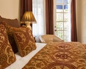 Close up of bed with rich red and gold pillows and bedspread, bedside lamp, leather chair and French doors