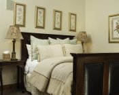 Dark wood and leather bed with ivory comforter, light green and ivory pillows, gold-framed pictures, and nightstands with lamps