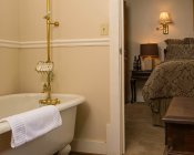 Clawfoot tub with brass faucet, ivory walls, bed in the background with brown and black comforter and dimly lit sconce