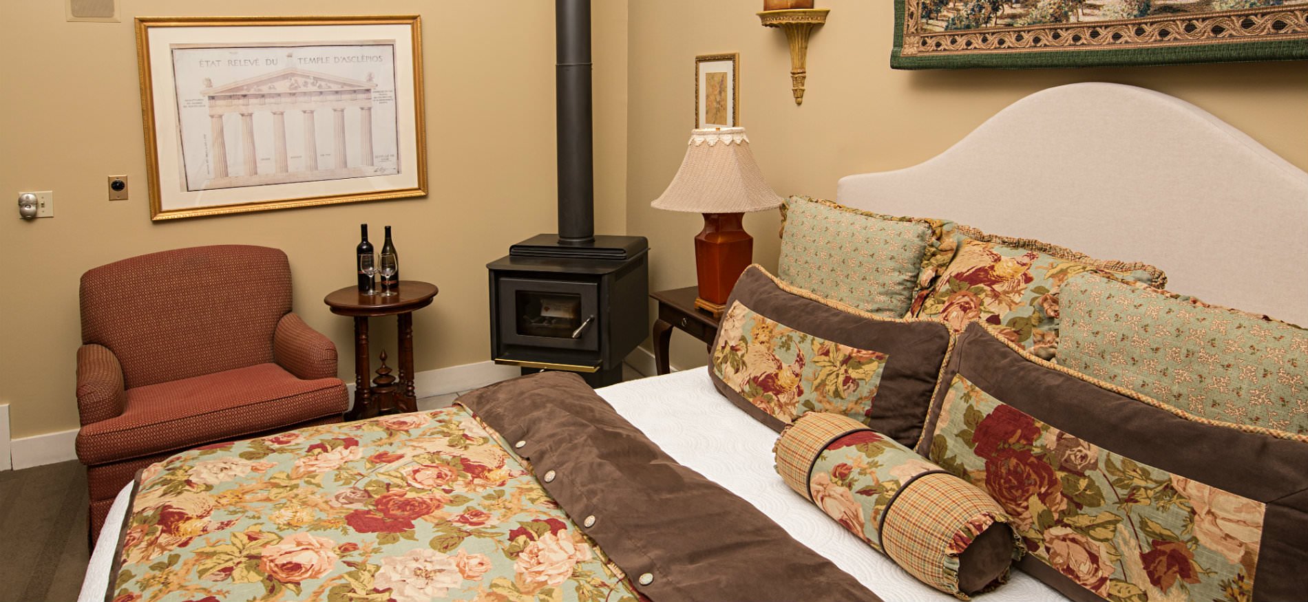 Bed with red, white, green, blue and brown floral comforter, tan walls, red chair with small table and wood stove