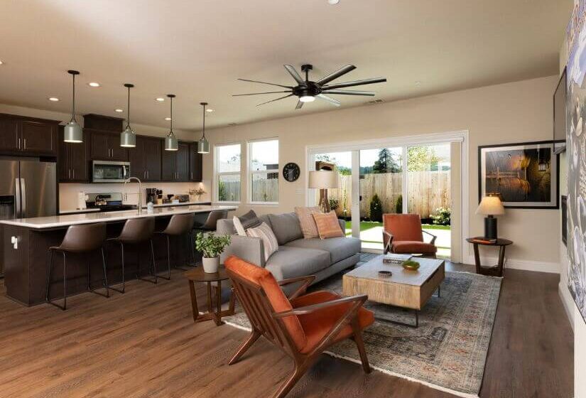 Living room with grey couch & orange chairs facing wall with tv mounted, kitchen with stainless steel appliance and dark cabinets plus island with bar chairs, sliding door to fenced yard.