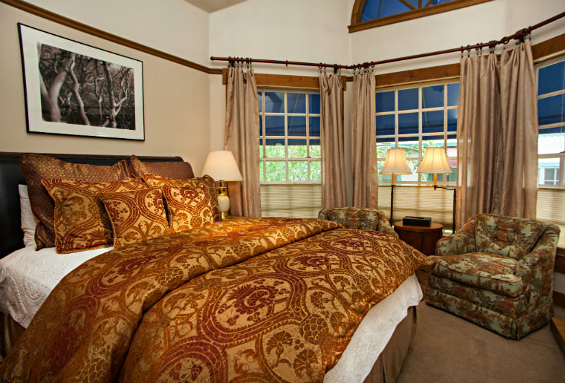 Bedroom with bay window, bed covered in red and gold pillows and comforter, two chairs and small table